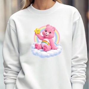 May include: White crewneck sweatshirt featuring a pink Care Bear character holding a yellow star. The bear sits on a cloud in front of a rainbow. The design is centered on the front of the sweatshirt.