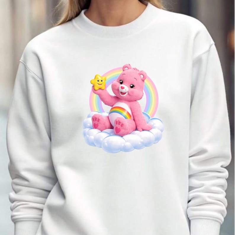 May include: White crewneck sweatshirt featuring a pink Care Bear character holding a yellow star. The bear sits on a cloud in front of a rainbow. The design is centered on the front of the sweatshirt.