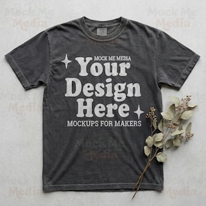 May include: Dark gray t-shirt with the text "Your Design Here" in white, along with the words "Mockups for Makers." A sprig of dried leaves rests on the right side of the shirt. The shirt is a mockup for makers.