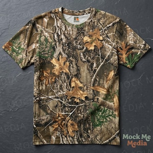 May include: Short-sleeved camouflage t-shirt with a crew neck. The shirt features a realistic forest pattern with brown, green, and tan hues, including leaves and branches. The Russell Athletic logo is visible on the neckline.