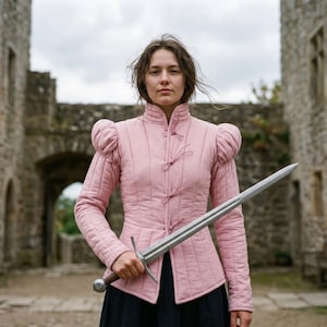 Peut inclure: Veste matelassée rose avec un col montant et des manches bouffantes. La veste a des attaches décoratives sur le devant. Une longue épée argentée est tenue devant la veste. Le fond présente un château en pierre.