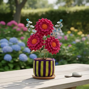 Pode incluir: Arranjo floral de crochê feito à mão. Apresenta três dalias magenta vibrantes com centros laranja, colocadas num vaso com riscas verticais amarelas e roxas. Detalhes azuis e brancos de crochê complementam o conjunto, sobre uma mesa de madeira.