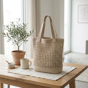 Könnte beinhalten: Beige gehäkelte Tragetasche mit offenem Gewebe und zwei Henkeln. Die Tasche steht auf einem Holztisch mit einer kleinen Topfpflanze, einer Tasse und einer kleinen Schale. Die Tasche hat einen rechteckigen Boden und ein strukturiertes, offenes Muster.