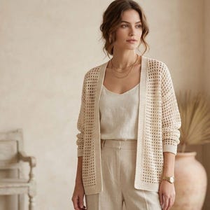 May include: A cream-colored, open-weave cardigan with long sleeves. The cardigan is worn over a cream-colored camisole and paired with matching pants. The outfit is accessorized with a gold watch and layered necklaces. The overall style is casual and comfortable.
