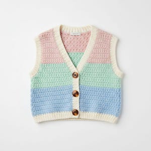Puede incluir: Un chaleco de crochet en colores pastel con cuello en V y tres botones marrones. El chaleco presenta rayas horizontales rosas, verdes y azules, bordeadas por un ribete color crema. La prenda sin mangas es una pieza acogedora y hecha a mano.