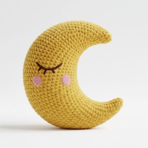 Könnte beinhalten: Ein gelbes gehäkeltes Halbmondkissen mit einem schläfrigen Gesicht. Das Kissen hat gestickte braune Wimpern und zwei rosa Wangenpunkte, die ein skurriles Design schaffen. Ideal für ein Kinderzimmer.