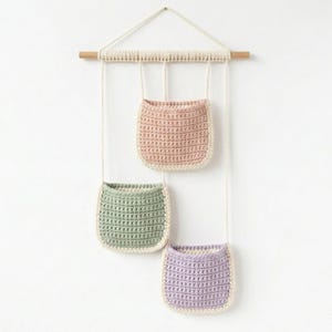 May include: A wall hanging with three crocheted pockets in pastel colors: pink, green, and purple. The pockets are suspended from a wooden dowel and rope, creating a decorative storage solution. The pockets have a cream-colored border.