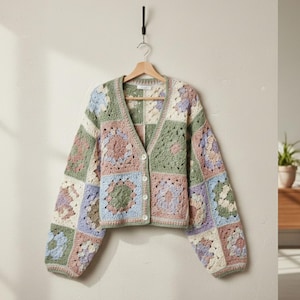 Könnte beinhalten: Ein mehrfarbiger Häkelcardigan mit V-Ausschnitt und Knopfverschluss. Der Cardigan hat ein Patchwork-Design mit Quadraten in Pastelltönen von Rosa, Blau, Grün und Creme. Er hängt an einem Holzbügel vor einer neutralen Wand.