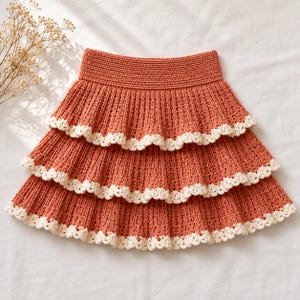 May include: A rust-colored crochet skirt featuring three tiered layers with a ribbed texture. Each tier is finished with a delicate, scalloped cream-colored crochet trim along the bottom edge, and the skirt has a wide, solid crochet waistband.