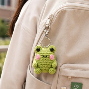 Può includere: Un portachiavi a forma di rana verde all'uncinetto con occhi neri e guance rosa è appeso a una clip argentata su uno zaino beige. La rana ha una bocca sorridente ed è fatta di filato. Lo zaino ha una cerniera.