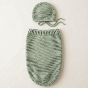 May include: A sage green crochet baby cocoon and matching bonnet set. The cocoon is a long, cylindrical shape with a ribbed top. The bonnet has a rounded crown and ties. The set is made of soft, textured yarn.