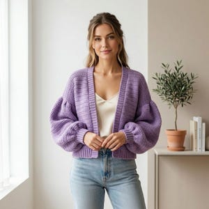 May include: A lilac-colored, open-front cardigan with long, puffy sleeves. The knitted sweater is worn over a cream-colored top and paired with light blue jeans. The cardigan is a soft, textured knit, perfect for layering.