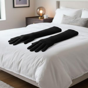 May include: Two long, black knitted gloves are laid out on a white bed. The gloves have a textured design and extend past the wrist. The bed is made with a white duvet cover and pillows, with a bedside table and lamp in the background.