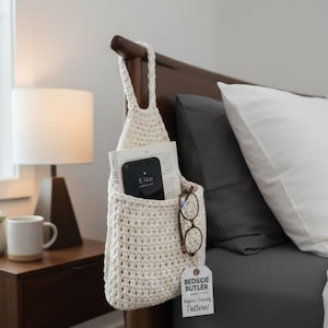 May include: A cream-colored, crocheted bedside organizer hanging from a wooden headboard. The organizer holds a book, a smartphone displaying 6:14 AM, and eyeglasses. A tag reads "Bedside Butler Beginners Friendly Pattern!"