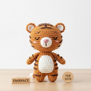 Puede incluir: Una figura de tigre de ganchillo en tonos naranja y marrón, con vientre y detalles blancos. El tigre tiene los ojos cerrados y una nariz rosa. Un bloque de madera con la palabra "EMBRACE" y otro con "FEB 2026" están en primer plano.