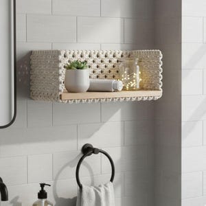 Puede incluir: Estante de macramé crema con base de madera, que contiene una pequeña planta en maceta, una toalla enrollada y un tarro con luces de hadas. El estante está montado en una pared de azulejos blancos. También se ven un toallero negro y un espejo.