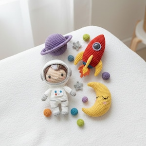 May include: A collection of crocheted space-themed toys, including an astronaut, a rocket, a planet, a crescent moon, and stars. The astronaut is wearing a white spacesuit. The rocket is red with yellow flames. The planet is purple, and the moon is yellow.