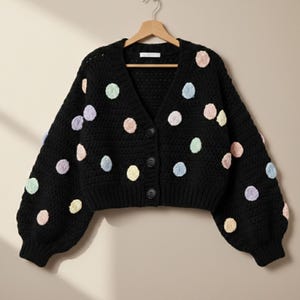 May include: A black, button-down cardigan sweater with a V-neck and long sleeves. The sweater is adorned with colorful, pastel-colored polka dots. The sweater is hanging on a wooden hanger.