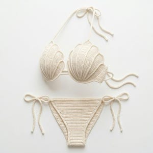 May include: Cream-colored crochet bikini set. The top features shell-shaped cups with tie straps. The bottoms are a classic bikini cut with tie sides. The set is made of a soft, textured yarn, perfect for beachwear or swimwear.