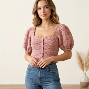Pode incluir: Top de crochê rosa empoeirado com decote quadrado e mangas curtas bufantes. O top apresenta uma borda ondulada e botões de madeira na frente. A modelo está usando jeans azuis. Peça de roupa feita à mão.