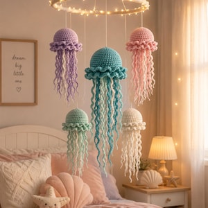 May include: A hanging mobile featuring five crocheted jellyfish in shades of lavender, teal, pink, mint green, and cream. The jellyfish are suspended from a circular frame wrapped in light-colored yarn and illuminated by small, warm white LED lights.