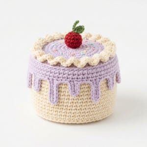 May include: A crocheted cake with a cream-colored base and a light purple top. The top has a cream-colored scalloped edge and is decorated with sprinkles and a red cherry. The purple icing drips down the sides.