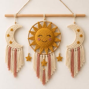 Op de afbeelding: Een handgemaakte wandhanger met een lachende zon en twee halve manen. De zon is geel met een lachend gezicht en de manen zijn crèmekleurig met sterren. De hanger bevat kwasten in crème en roze, en houten kralen.