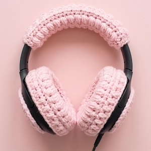 May include: A pair of black headphones with light pink crocheted ear covers and headband cover. The crochet work has a textured appearance, and the background is a matching light pink.