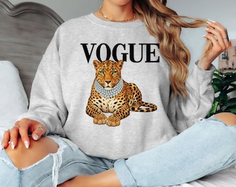 May include: A light gray sweatshirt featuring the word "VOGUE" in black capital letters above a leopard illustration. The leopard is wearing a pearl necklace. The sweatshirt is a casual style.