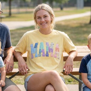 May include: A woman wearing a yellow t-shirt with the word "MAMA" in patchwork letters. The letters are made of various patterned fabrics in shades of blue, purple, and pink. She is sitting on a wooden bench outdoors.