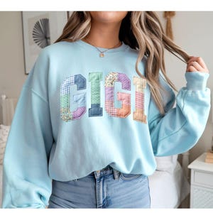 Può includere: Una felpa azzurra con la scritta "GIGI" a lettere patchwork. Le lettere sono realizzate con vari tessuti fantasia nei toni del blu, verde, viola e pesca. La felpa ha maniche lunghe e girocollo.