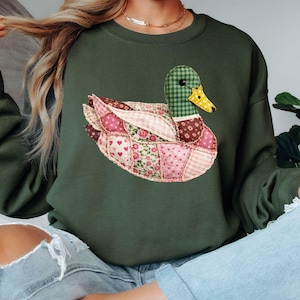 May include: An olive green sweatshirt with a patchwork duck design. The duck has a green and yellow checkered head, a yellow beak, and a body made of pink and floral patterned fabrics. The sweatshirt is a casual style.