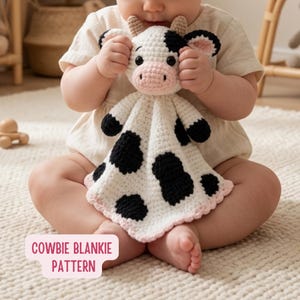 May include: A crochet cow security blanket featuring a white body with black spots, a pink snout, and small horns. The toy has black eyes and a light pink scalloped edge. A pink label at the bottom displays the text COWIE BLANKIE PATTERN.