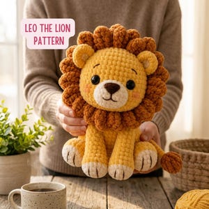 May include: A crocheted lion plush toy with a yellow body, a brown textured mane, black safety eyes, and a brown nose. The toy features light pink blush on the cheeks and white paws with brown stitched details. Text reads: LEO THE LION PATTERN.