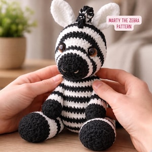 Może przedstawiać: Ręcznie robiona szydełkowa pluszowa zebra z czarno-białymi paskami, dużymi brązowymi oczami i białymi uszami. Widoczny jest tekst "Marty the Zebra Pattern". Zebra siedzi na drewnianej powierzchni.