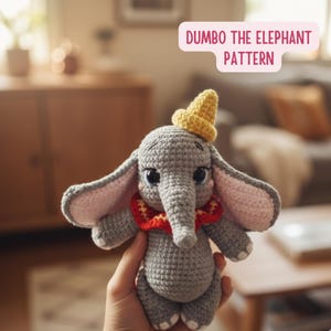 Puede incluir: Un juguete de elefante gris tejido a ganchillo, con orejas grandes, un sombrero amarillo y un cuello rojo. El juguete está hecho a mano y presenta el texto "Dumbo the Elephant Pattern" en una burbuja rosa.