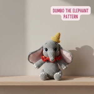 Puede incluir: Un juguete de elefante gris tejido con grandes orejas, un sombrero amarillo y un cuello rojo. El elefante tiene ojos azules y está sentado en un estante de madera. El texto "DUMBO THE ELEPHANT PATTERN" está en una burbuja rosa encima del juguete.