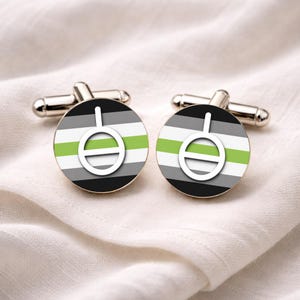 Può includere: Un paio di gemelli argentati con un design circolare raffigurante la bandiera dell'orgoglio aromantico. Il design della bandiera include strisce orizzontali nere, grigie, bianche e verdi, con un simbolo bianco al centro.