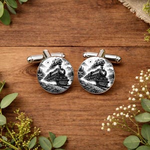 May include: A pair of silver cufflinks with a black and white train design. Each cufflink features a detailed illustration of a vintage steam train, set against a backdrop of trees and a mountain range. The cufflinks have a classic, round shape.