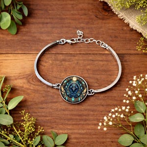 May include: Silver-toned bracelet with a circular pendant featuring a detailed design in shades of blue, teal, and gold. The pendant has a central bird-like figure and is set against a starry background. The bracelet is on a wooden surface.