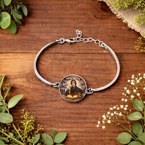 Peut inclure: Un bracelet argenté avec un médaillon rond orné d'une illustration d'un homme couronné tenant une épée et un livre. Le mot "MACBETH" est imprimé au-dessus. Le bracelet est posé sur une surface en bois.