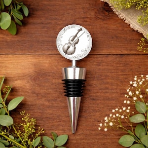 Può includere: Un tappo per bottiglia di vino argentato con una guarnizione in gomma nera e una punta conica. Il tappo presenta un design circolare con un'illustrazione dettagliata di un violino e note musicali. Lo sfondo è una superficie di legno calda.
