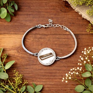 May include: Silver-toned bracelet with a round charm featuring a keyboard design. The charm is white with a brown keyboard illustration and musical notes. The bracelet is set against a dark wood surface with greenery.