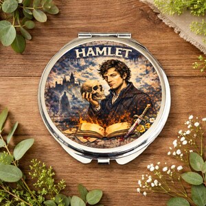 Op de afbeelding: Ronde, zilveren compacte spiegel met een illustratie uit Hamlet. De afbeelding toont een man die een schedel vasthoudt, met het woord "HAMLET" bovenaan. Een brandend boek en een zwaard zijn ook zichtbaar.