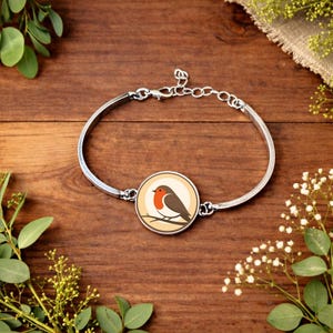 Könnte beinhalten: Silbernes Armreif-Armband mit einem runden Anhänger mit einem Rotkehlchen-Design. Der Vogel hat eine rote Brust und einen braunen Körper und sitzt auf einem Ast. Das Armband liegt auf einem Holzgrund mit Grünpflanzen.