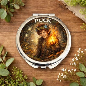 Op de afbeelding: Een zilveren compacte spiegel met het woord "PUCK" in witte letters. De afbeelding van de spiegel toont een lachende persoon met een bloemenkrans die een vuurbal vasthoudt. De achtergrond toont een bosscène met paddenstoelen en gloeiende lichten.
