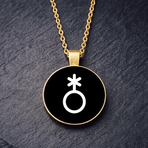 May include: A gold-colored necklace with a round pendant featuring a white symbol on a black background. The chain and pendant holder are gold, and the symbol is a circle with a cross above it. The necklace is displayed on a dark, textured surface.