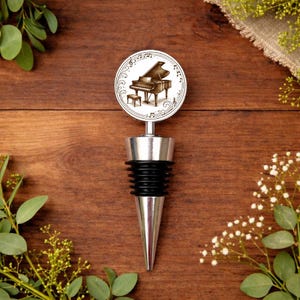 May include: A silver and black wine bottle stopper with a piano design. The round top features a sepia-toned illustration of a grand piano and music notes. The stopper has a black rubber seal and a conical silver base. The background is a wooden surface.