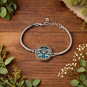 Pode incluir: Uma pulseira prateada com um pendente circular com um design de tridente dourado sobre um padrão de ondas azuis e brancas. A pulseira é exibida em uma superfície de madeira, rodeada por vegetação.