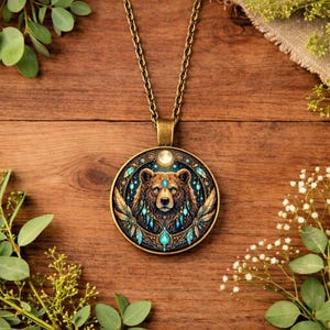 Könnte beinhalten: Halskette mit bronzefarbenem Anhänger mit detaillierter Bärenillustration. Der runde Anhänger hat türkisfarbene und goldene Akzente und eine Kette. Die Halskette liegt auf einem hölzernen Hintergrund mit Grün.